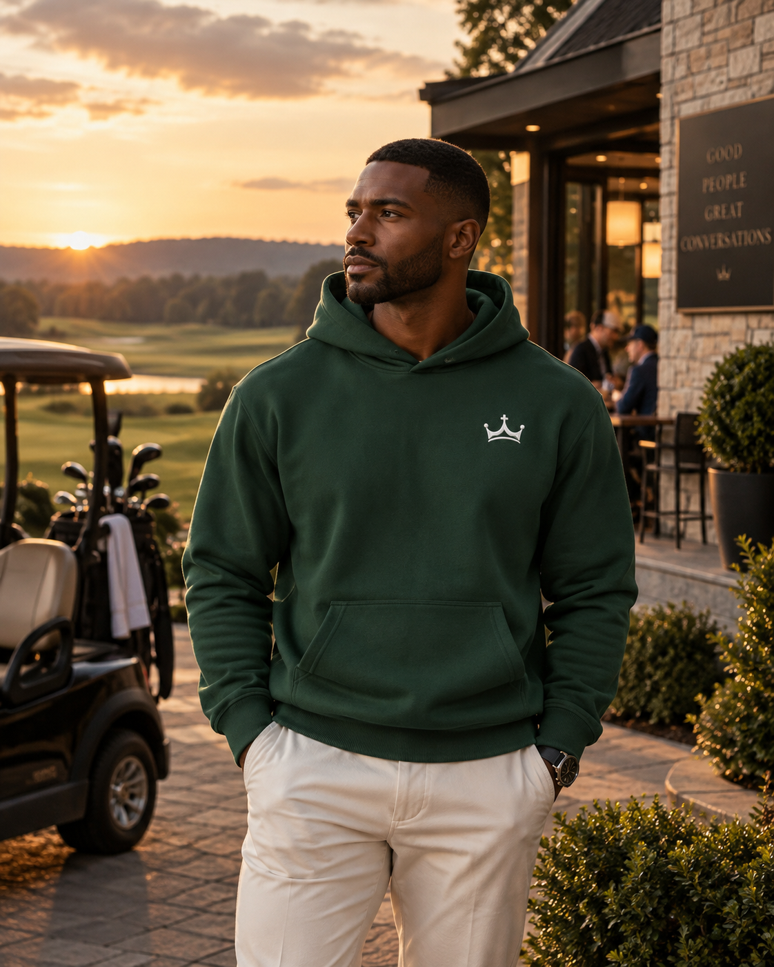 Man in a green Crown Hoodie standing outside the clubhouse at sunset, golf cart and course in the background