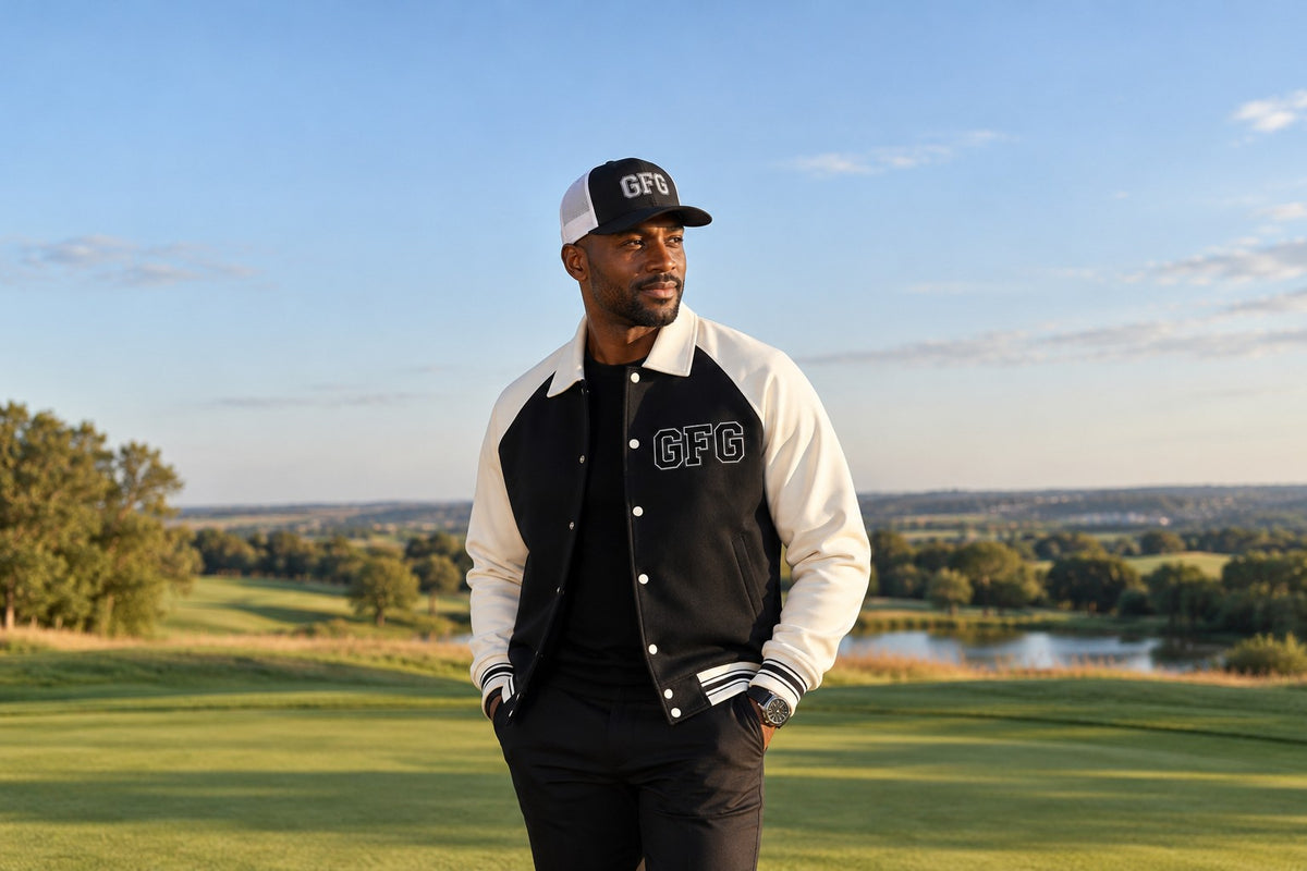 Man in a black and cream GFG varsity jacket and trucker cap on a golf fairway at golden hour